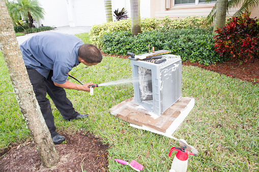 An air conditioner repairman washing the coils from an air conditioning unit. He is working on the front lawn of a home.